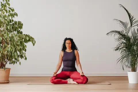 Frau praktiziert Yoga zu Hause im Lotussitz, umgeben von Pflanzen.