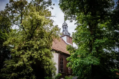 Stary kościół w Miejskiej Górce, otoczony bujną zielenią drzew, z charakterystyczną wieżą i drewnianą elewacją.