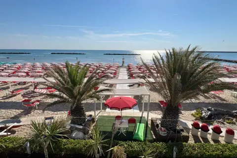 Spiaggia di Porto San Giorgio mare con file ordinate di ombrelloni rossi e bianchi, lettini, palme e il mare azzurro sotto un cielo limpido.