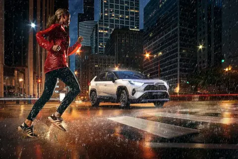 Kobieta biegnie w deszczu po mieście, obok niej jedzie biała Toyota RAV4 Hybrid. Zastanawiasz się, ile pali Toyota RAV4?
