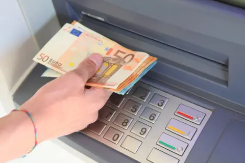 Dłoń z banknotami 50 euro wkłada pieniądze do bankomatu.
