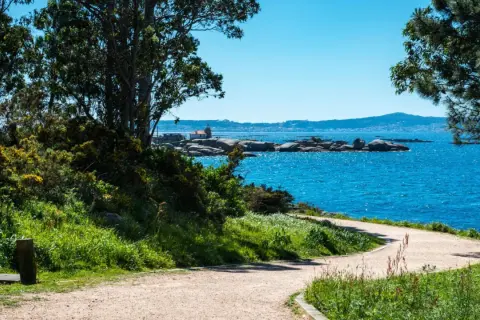 Sendero costero en la Illa de Arousa con mar azul brillante, rocas y un pequeño faro.