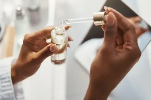 Dłonie trzymają buteleczkę z serum. Kropla z pipety spada do butelki, być może z kwasem ferulowym, dla zdrowej skóry.