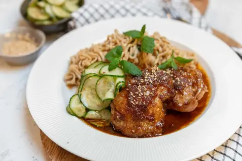 Två saftiga bitar sticky chicken med sesamfrön, serverade med nudlar och gurksallad.