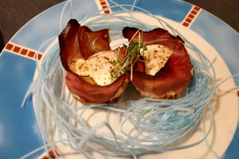 Zwei Eier mit Speck, kunstvoll in einem Nest aus blauen Fäden auf einem Teller angerichtet.
