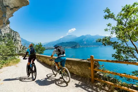 Ciclisti su una strada panoramica lungo il Lago di Garda, con montagne sullo sfondo.