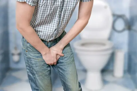Mężczyzna w dżinsach i koszuli w kratę trzyma ręce w kroku, wskazując na problem z pęcherzem neurogennym. W tle widoczna toaleta.