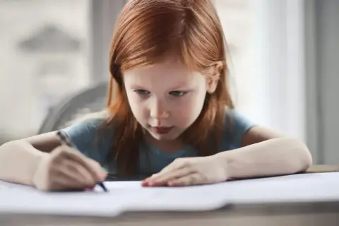 Ruda dziewczynka skupiona na swoich pracach domowych, pisze coś na kartce.