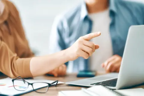 Dłoń wskazująca palcem na ekran laptopa, obok okulary i wykresy.