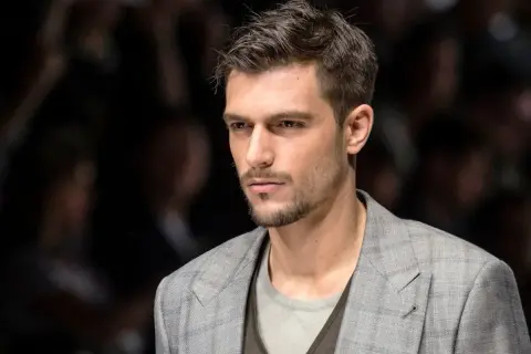 Uomo con capelli spettinati corti, sguardo intenso, indossa una giacca a quadri grigia.