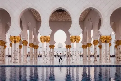Un hombre camina bajo arcos blancos y dorados en Abu Dabi, reflejándose en el agua. Un visado Abu Dabi es el primer paso para disfrutar de esta maravilla.