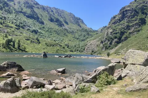 Lac de montagne entouré de montagnes verdoyantes et de rochers, sous un ciel bleu clair.