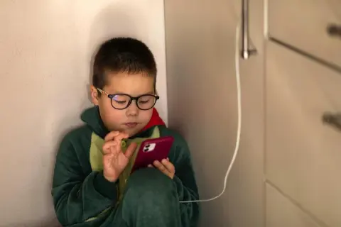 Chłopiec w okularach i piżamie z dinozaurem skupiony na telefonie. Czas na ekranie to wyzwanie, ale warto pamiętać o ograniczeniu czasu na telefonie.