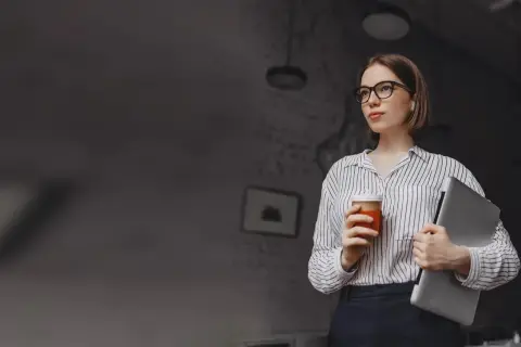 Kobieta w okularach trzymająca kawę i laptopa.