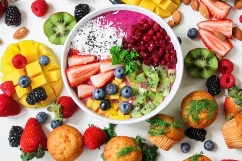 Kolorowa miska smoothie z owocami: truskawki, kiwi, mango, jagody, maliny, jeżyny, granat. Obok muffinki i migdały. Zdrowe produkty, które mogą zawierać histaminę.