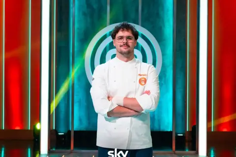 Matteo, vincitore MasterChef 15, posa con la sua divisa da chef, braccia conserte, sullo sfondo del logo del programma.