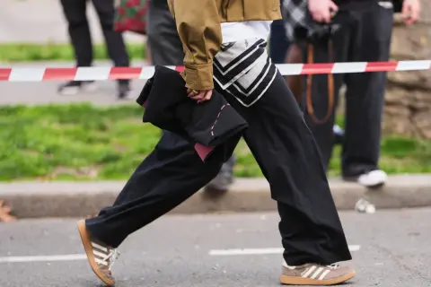 Persona con un outfit casual, pantaloni neri ampi, giacca color kaki e scarpe da ginnastica Adidas marroni.