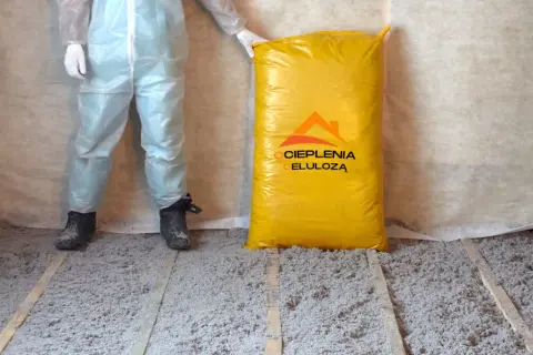 Osoba w kombinezonie ochronnym stoi obok worka z materiałem do ocieplenia celulozą. Podłoga pokryta jest warstwą izolacji.