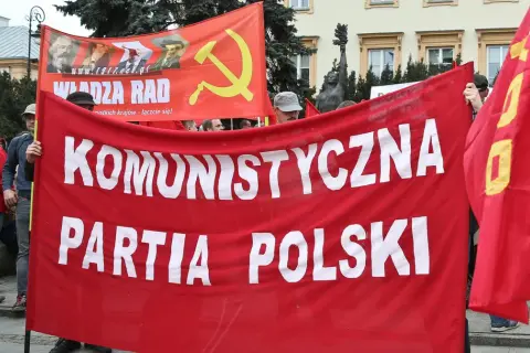 Ludzie niosą czerwony transparent "KOMUNISTYCZNA PARTIA POLSKI" i flagę z sierpem i młotem. To manifestacja poparcia dla socjalizmu.