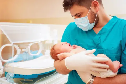 Lekarz w maseczce i rękawiczkach trzyma noworodka, troskliwa neonatologia.