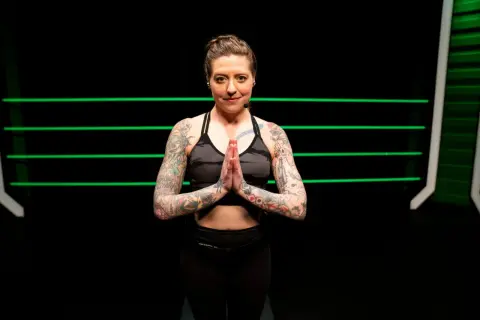 A tattooed woman in athletic wear holds her hands in prayer pose, ready for yoga for boxers.