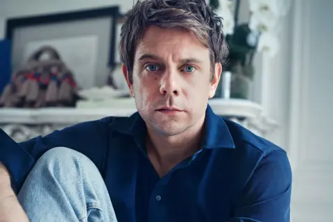 Ritratto di Jonathan Anderson, con occhi azzurri penetranti, indossa una camicia blu e jeans.