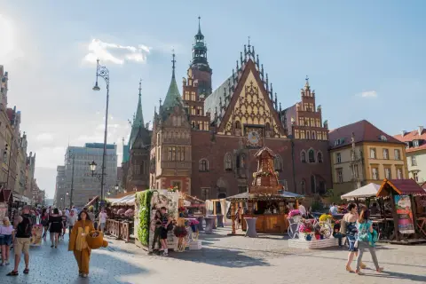Tętniący życiem wrocławski Rynek, z Ratuszem w tle. Ludzie spacerują wśród kramów, a ktoś w stroju misia dodaje uroku temu miejscu.