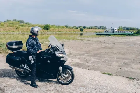 Motocyklista w kasku i kurtce stoi obok czarnego motocykla nad kanałem z łodzią w tle.