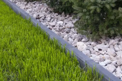 Zgrabna rabata z zieloną trawą i kamieniami, oddzielona betonowym obrzeżem. Suchy beton pod obrzeża zapewnia stabilność.