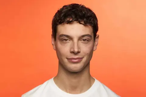 Hombre joven con cabello castaño rizado, luciendo una permanente ondulada. Sonríe levemente ante un fondo naranja vibrante.