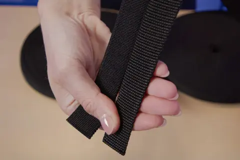 Dłoń trzyma czarną taśmę parcianą, zwoje materiału w tle.