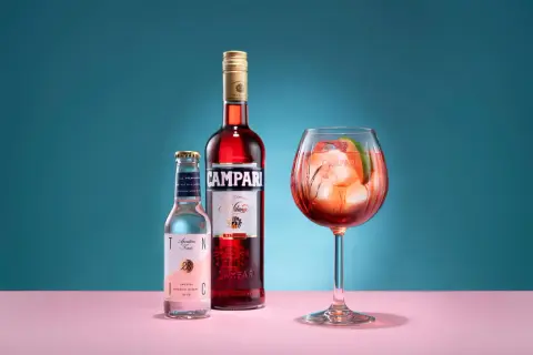 En Campari Spritz med is och lime i ett stort glas. Bredvid står en flaska Campari och en flaska tonic.