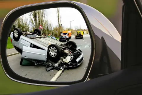 Auto w leasingu leży na dachu po wypadku. Widok w lusterku bocznym pokazuje szkody z OC sprawcy i pomoc drogową na drodze.