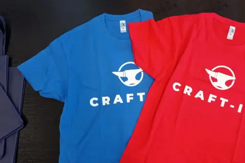 Niebieska i czerwona koszulka z logo "CRAFT-IT" i kowadłem. To przykład, jak sitodruk co to za technika, pozwala tworzyć trwałe nadruki na odzieży.