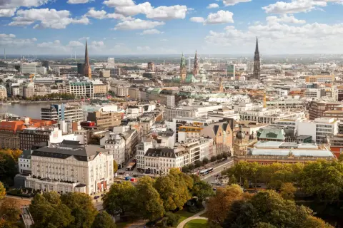 Blick von einer **Aussichtsplattform Hamburg** über die Stadt mit ihren Kirchtürmen, dem Hafen und grünen Bäumen im Vordergrund.