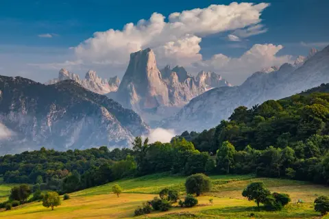 Majestuosas montañas de los Picos de Europa se alzan tras un prado verde y un bosque frondoso, bajo un cielo azul con nubes algodonosas.