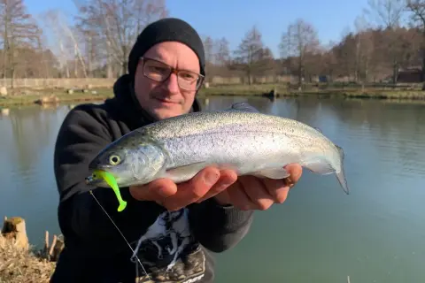 Glücklicher Angler hält eine frisch gefangene quellental forelle mit einem leuchtend grünen Köder im Maul.