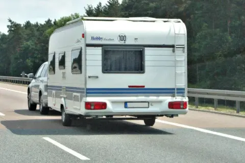 Biała przyczepa kempingowa Hobby jedzie autostradą. Kierowca z kod 96 prawo jazdy wiezie rodzinę na wakacje.
