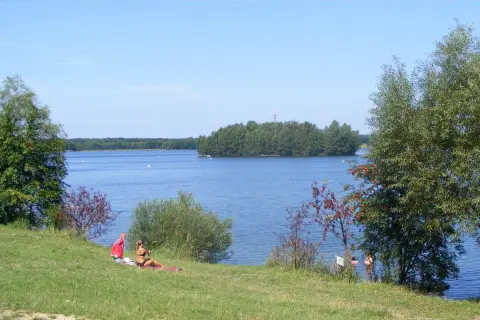 Słoneczny dzień nad jeziorem Chechło-Nakło. Kobieta odpoczywa na trawie, w tle błękitna woda i zielone wyspy.