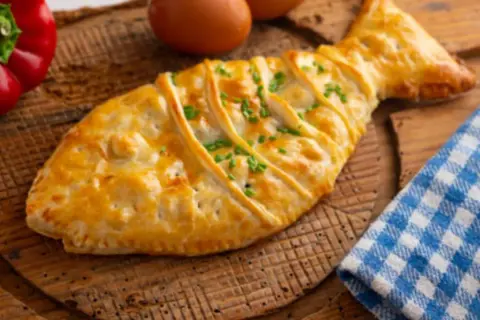 Empanada de bacalao con forma de pez, dorada y adornada con cebollino. Acompañada de pimiento rojo y huevos.