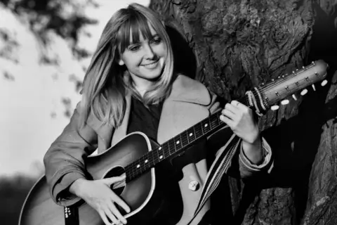 Maryla Rodowicz z gitarą, jakby śpiewała "Wielka woda", oparta o drzewo.
