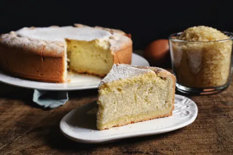 Una fetta di torta di riso, la ricetta originale, cosparsa di zucchero a velo, accanto alla torta intera e a una tazza di riso.