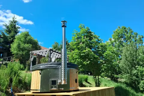 Drewniane jacuzzi w ziemi z metalowym kominem, otoczone zielenią i błękitnym niebem. Idealne miejsce na relaks.