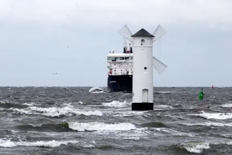 Biała latarnia morska w kształcie wiatraka na wzburzonym morzu, obok statku.