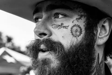Zbliżenie na twarz Post Malone'a w czerni i bieli, z tatuażem "Tired" na policzku. Fani w Polsce czekają na jego koncert.