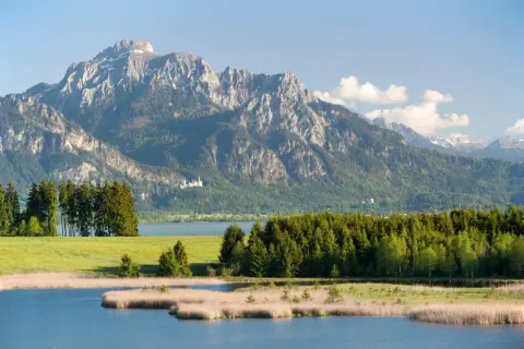 Majestätischer berg mit see und einer kleinen Burg im Hintergrund. Grüne Wiesen und Schilf am Ufer.