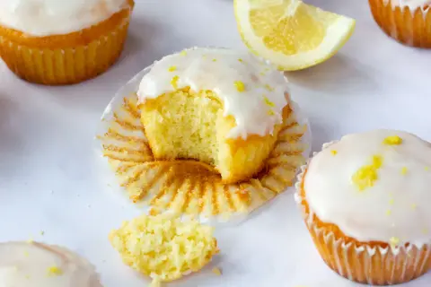 Saftige Zitronen-Magdalenas, eines davon angeknabbert, mit Zuckerguss und Zitronenschale bestreut. Ein perfektes magdalenas rezept für den Nachmittagstee.