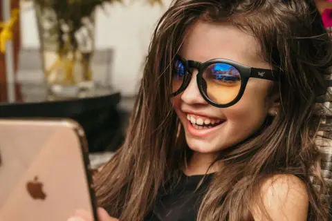 Uśmiechnięta dziewczynka w okularach z filtrem światła niebieskiego patrzy na telefon.