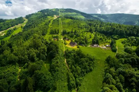 Widok z lotu ptaka na zielone zbocza Skrzycznego, z widocznymi trasami narciarskimi i szlakami pieszymi, domkami i kolejką linową.