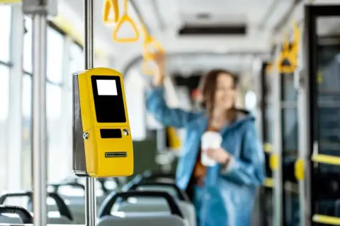 Żółty biletomat w autobusie, obok siedzeń. Kobieta w tle trzyma kubek. Pomyśl o zniżkach i ulgach na przejazdy.